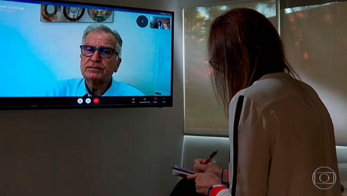A imagem mostra uma mulher de costas para a câmera, olhando para uma tv onde está transmitindo a câmera do Prof. Dr. Luiz Carneiro.