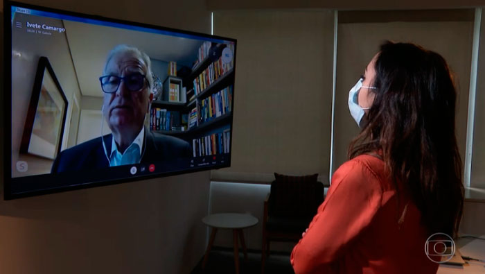 A imagem mostra uma mulher de costas para a câmera, na diagonal, olhando para um televisão. Nesta televisão, há uma chamada de vídeo com o Prof. Dr. Luiz Carneiro.