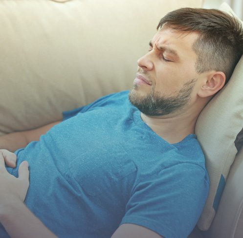 Ao fundo da imagem, há um homem deitado no sofá com as mãos no local no pâncreas e uma expressão de dor no rosto.