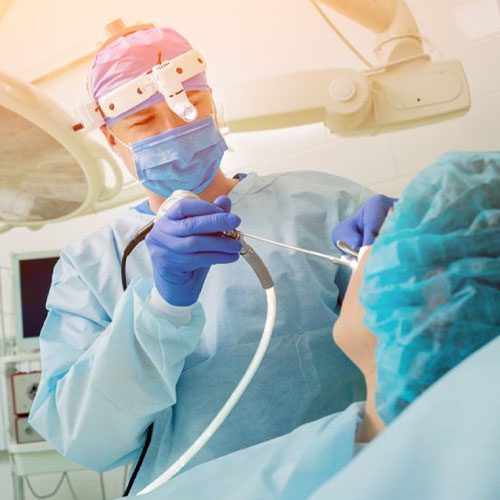 A imagem mostra uma paciente deitada na maca de um hospital (de costas para a câmera) e o doutor à sua frente com um equipamento em sua boca.
