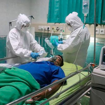 A imagem mostra dois médicos usando roupa completa de proteção contra doenças transmissíveis cuidando de um paciente.