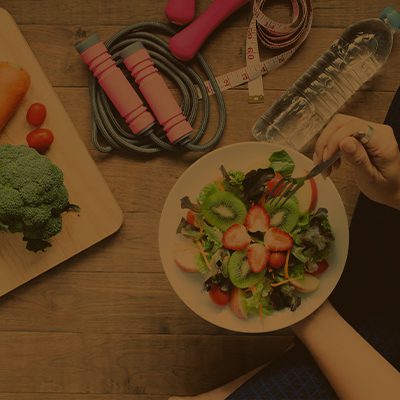 A imagem mostra mãos segurando um prato com frutas, uma tábua de cortar com vegetais e ao lado uma corda de pular, uma garrafa com água e uma fita métrica.