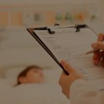 A imagem mostra uma pessoa segurando uma prancheta com uma folha de papel sulfite em uma mão e a outra está escrevendo algo com uma caneta. No fundo da imagem há uma pessoa deitada em uma cama de hospital.