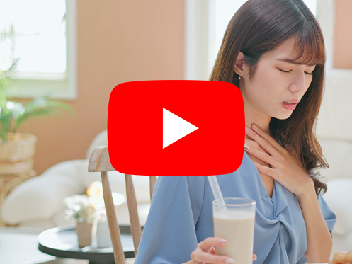 A imagem mostra uma mulher sentada em uma cadeira segurando um copo de leite enquanto está com a outra mão na garganta, como se estivesse sentindo algo.