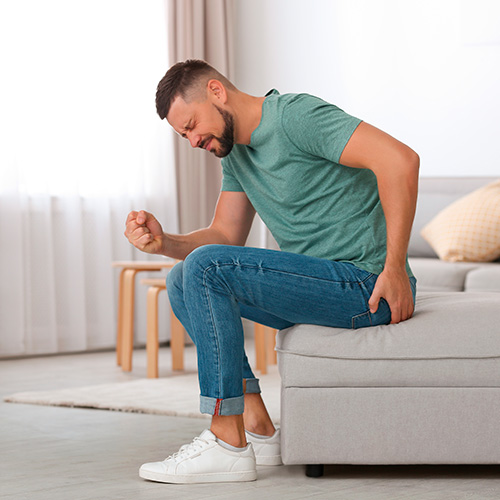 Homem sentado em sofá com expressão de dor, segurando a região anal, sugerindo desconforto relacionado a hemorroidas ou pós-operatório.