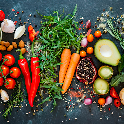 Variedade de alimentos frescos sobre superfície escura, incluindo cenouras, abacate, pimentões, tomates, alho, folhas verdes e especiarias.