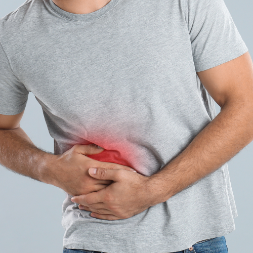 Homem com camiseta cinza sente dor abdominal, segurando a região lateral do abdômen com vermelhidão destacada.