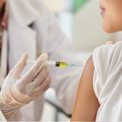Profissional de saúde aplicando uma injeção no braço de uma paciente, usando luvas e seringa com líquido amarelado.