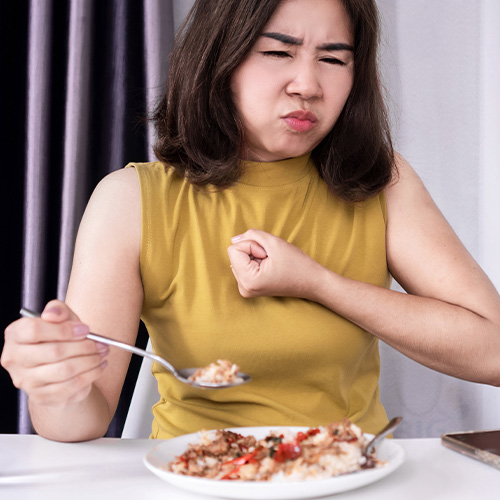 Mulher à mesa comendo e demonstrando desconforto no peito ao engolir, sugerindo dificuldade.