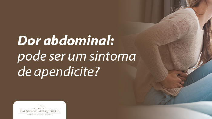 Mulher sentada em um sofá com as mãos sobre o lado direito do abdômen, demonstrando expressão de dor e desconforto, em ambiente interno iluminado pela luz da janela.